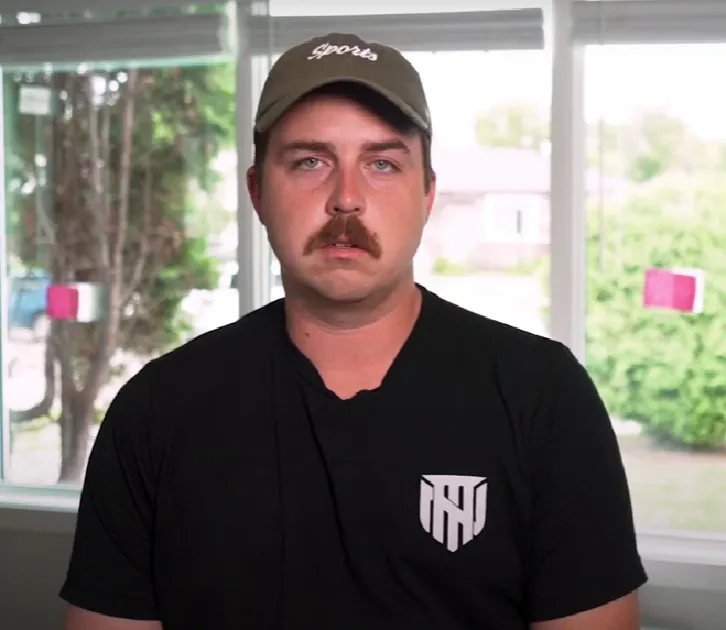 Man with a mustache wearing a black t-shirt and a green cap with the word 'sports' embroidered on it, sitting indoors in front of a window.