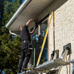 Technician installing or repairing a security camera on a house exterior near a window.
