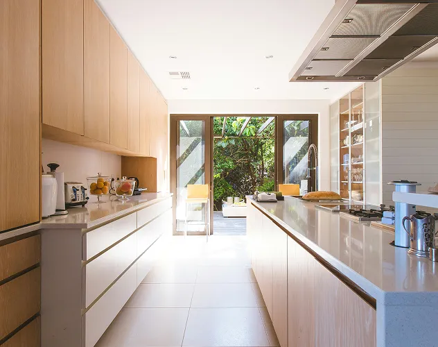 Bright modern kitchen with light wood cabinetry, white countertops, and large glass doors opening to an outdoor area with greenery.