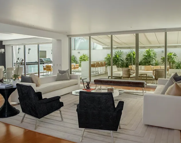 Modern living room with white sofas, two black armchairs, glass coffee table, and large sliding glass doors opening to an outdoor patio with seating and greenery.