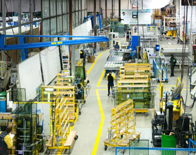 Industrial warehouse with workers and yellow-framed glass panels stored on wheeled racks along a marked central walkway.