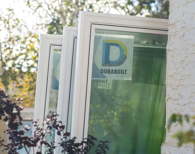 Three white-framed glass windows leaning against a textured white wall with a Durabuilt logo sticker on the nearest window.