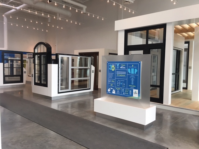 Modern living room with a gray chaise lounge near large windows showing outdoor trees, a floor lamp, and a TV on a stand with decorative items.