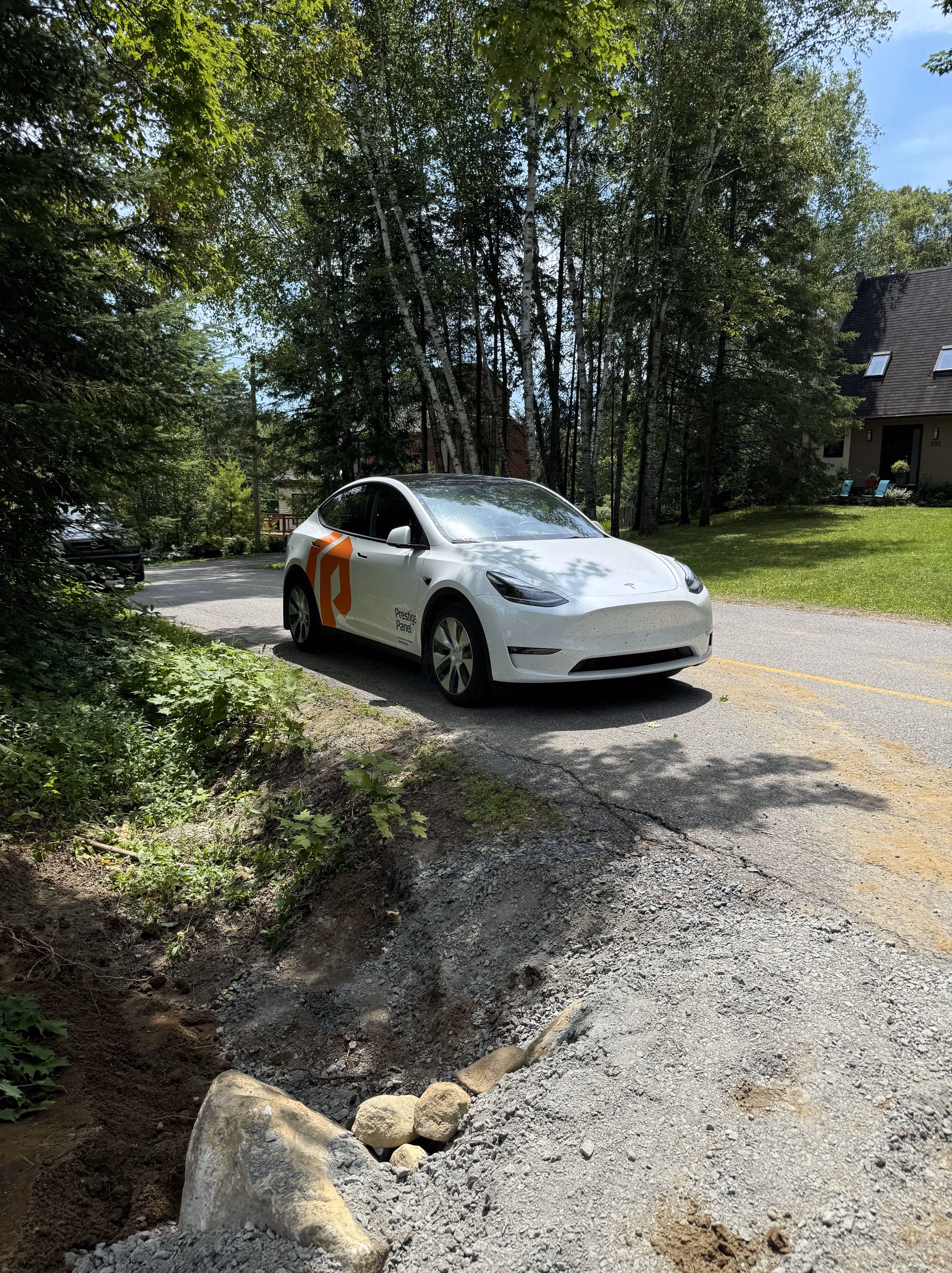 Voiture électrique blanche avec logo orange Prestige Panel sur une route entourée d'arbres et de maisons.