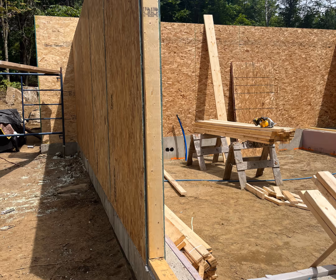 Chantier de construction avec murs en panneaux de bois osb, bûches empilées sur tréteaux et outils de menuiserie.