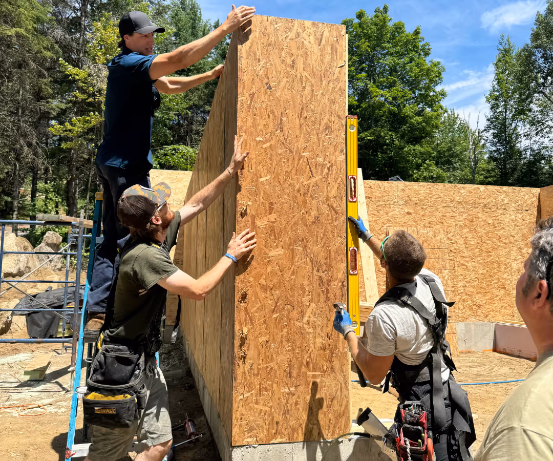 Quatre travailleurs de la construction installent un panneau en bois sur une fondation extérieure sous un ciel bleu.