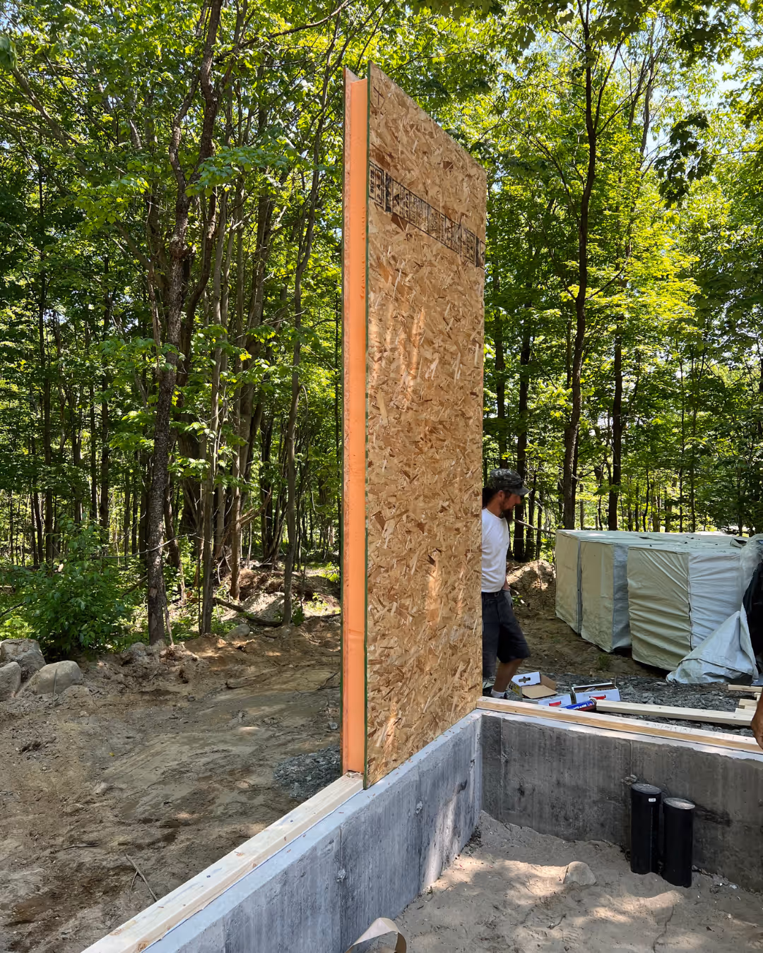 Un panneau isolant debout en construction sur une fondation en béton entourée d'arbres verts, avec un homme portant un chapeau debout à côté.