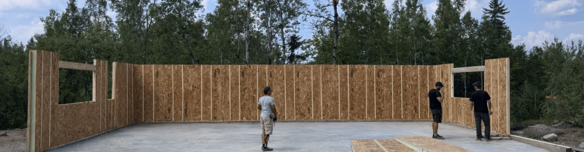 Trois hommes debout à l'intérieur d'une structure en construction avec des murs en panneau de bois entourée d'arbres.
