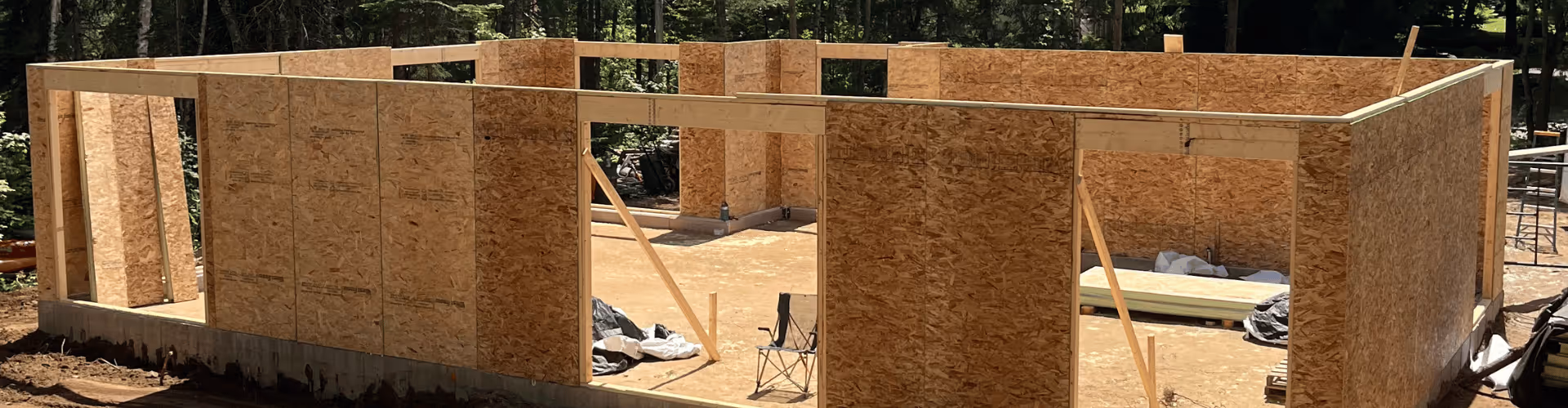 Structure en bois d'une maison en construction avec murs en panneaux OSB installés sur une dalle en béton entourée d'arbres.