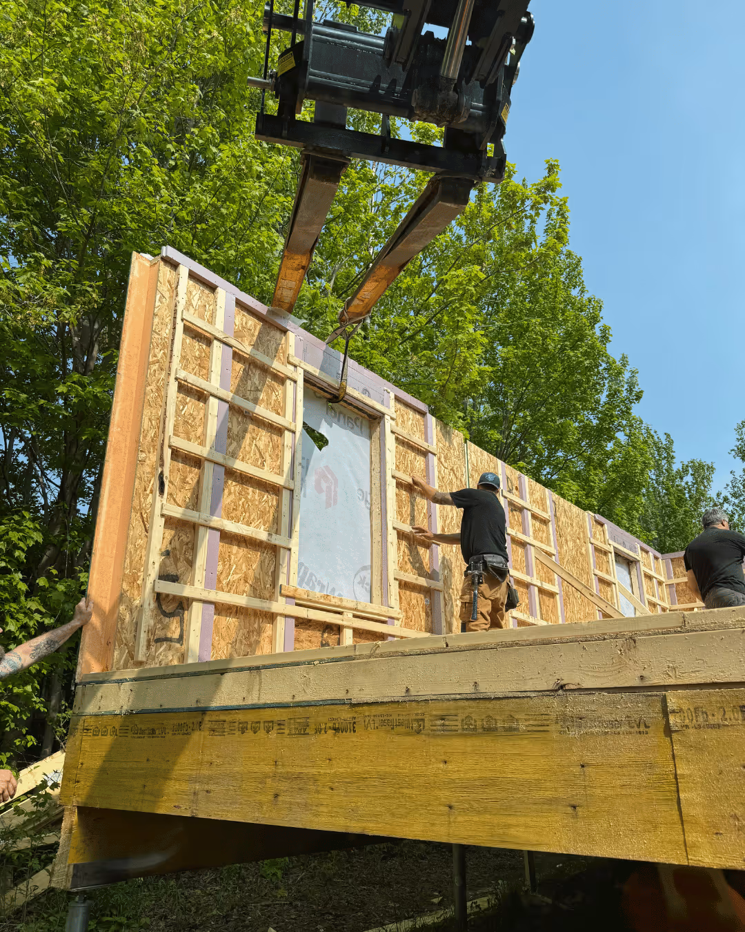Travailleurs en train d'installer un mur en bois préfabriqué sur une structure en bois extérieure entourée d'arbres verts par une journée ensoleillée.