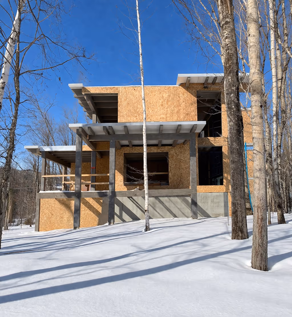 Maison en construction avec ossature en bois et béton entourée d'arbres en hiver sous un ciel bleu clair.