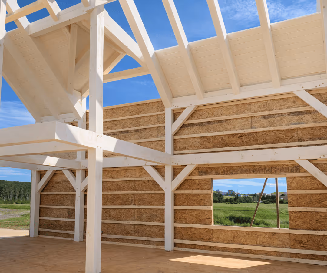 Structure en bois clair d'une maison en construction avec une fenêtre ouverte donnant sur un champ vert et un ciel bleu.
