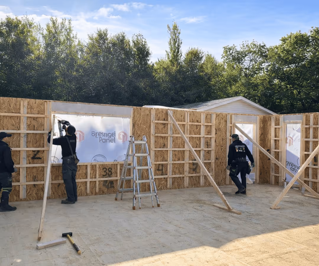 Deux ouvriers en construction installant des panneaux isolants sur une structure en bois partielle, avec un escabeau au centre et un marteau au sol, sous un ciel bleu avec des arbres en arrière-plan.