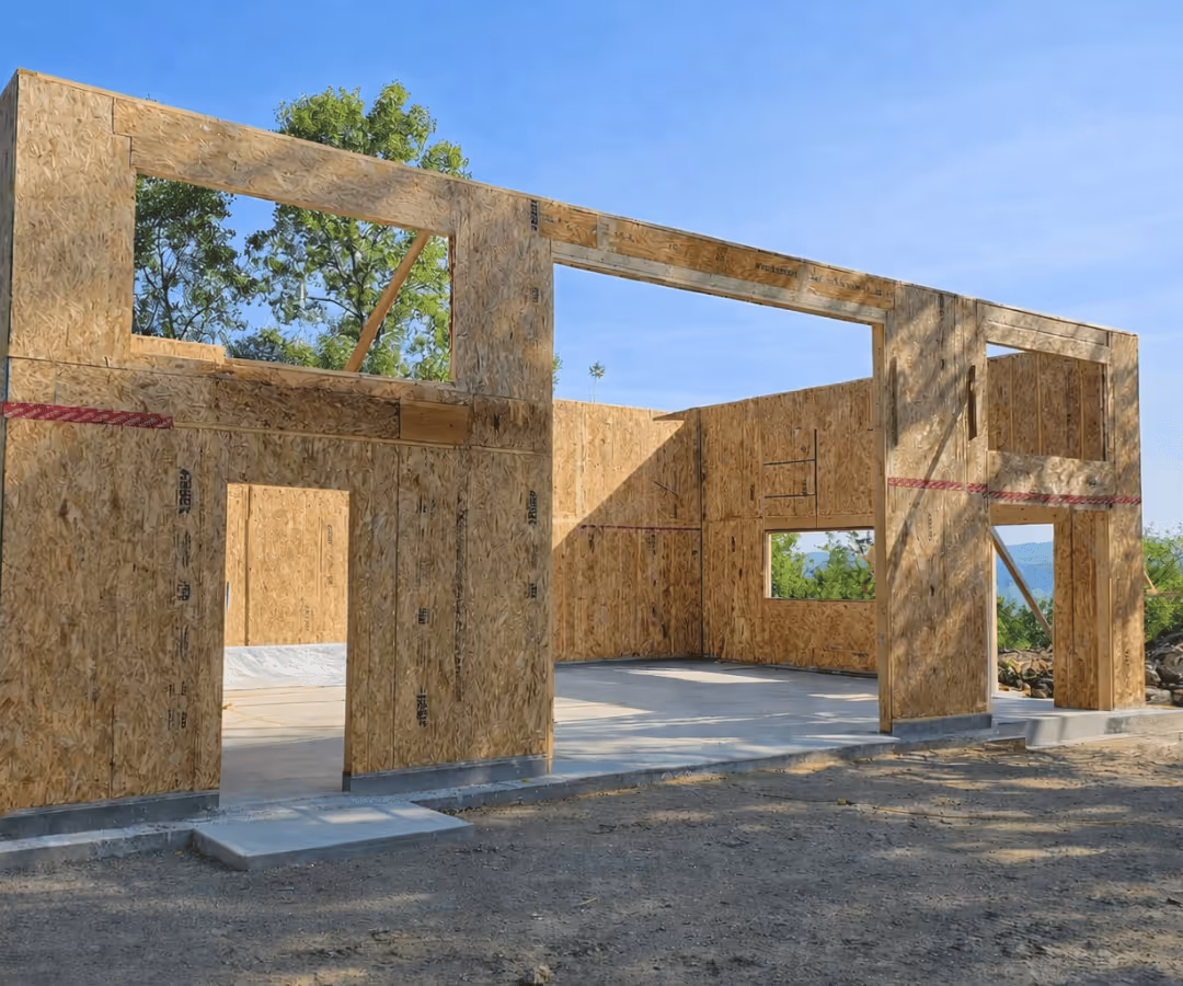 Structure en construction en panneaux de bois OSB avec ouvertures pour portes et fenêtres sur fond de ciel bleu et arbres verts.