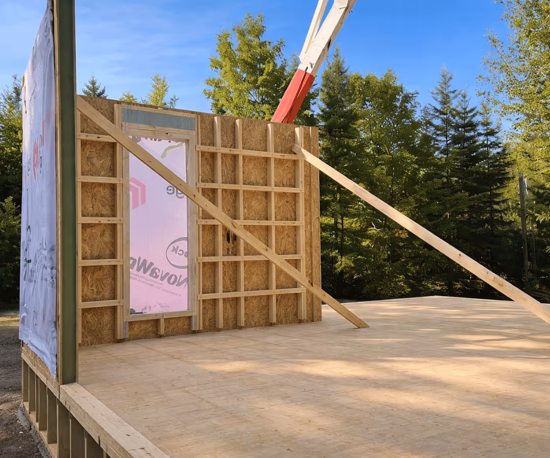 Structure en construction avec un mur partiel en bois et une base en contreplaqué dans un cadre boisé sous un ciel bleu.