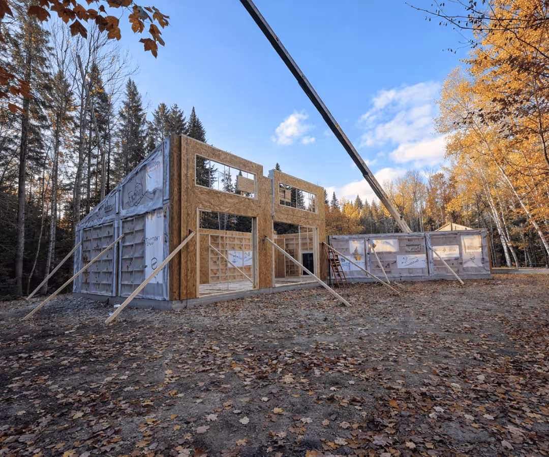 Construction d'une maison en bois avec des murs partiellement installés et un ciel bleu clair en arrière-plan, entourée d'arbres d'automne.