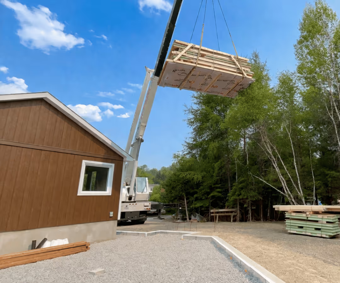 Grue soulevant un lot de matériaux de construction au-dessus d'un chantier près d'une maison en bois sous un ciel bleu avec quelques nuages.