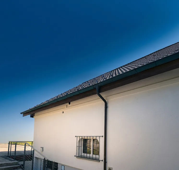 Hausfassade mit weit vorstehendem Dach und vergittertem Kellerfenster vor blauem Himmel