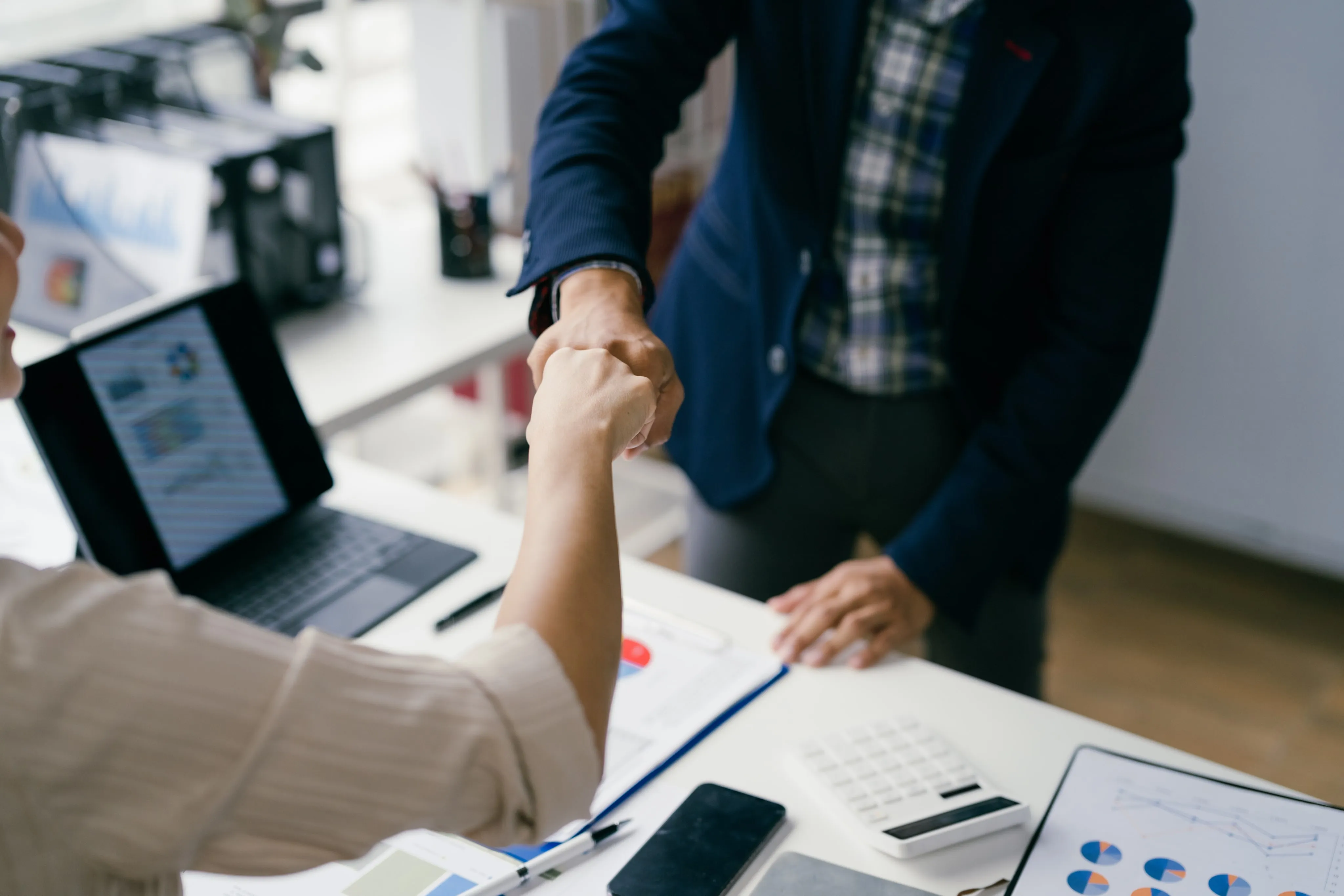 two businesspeople shaking hands over desk with documents and laptop








