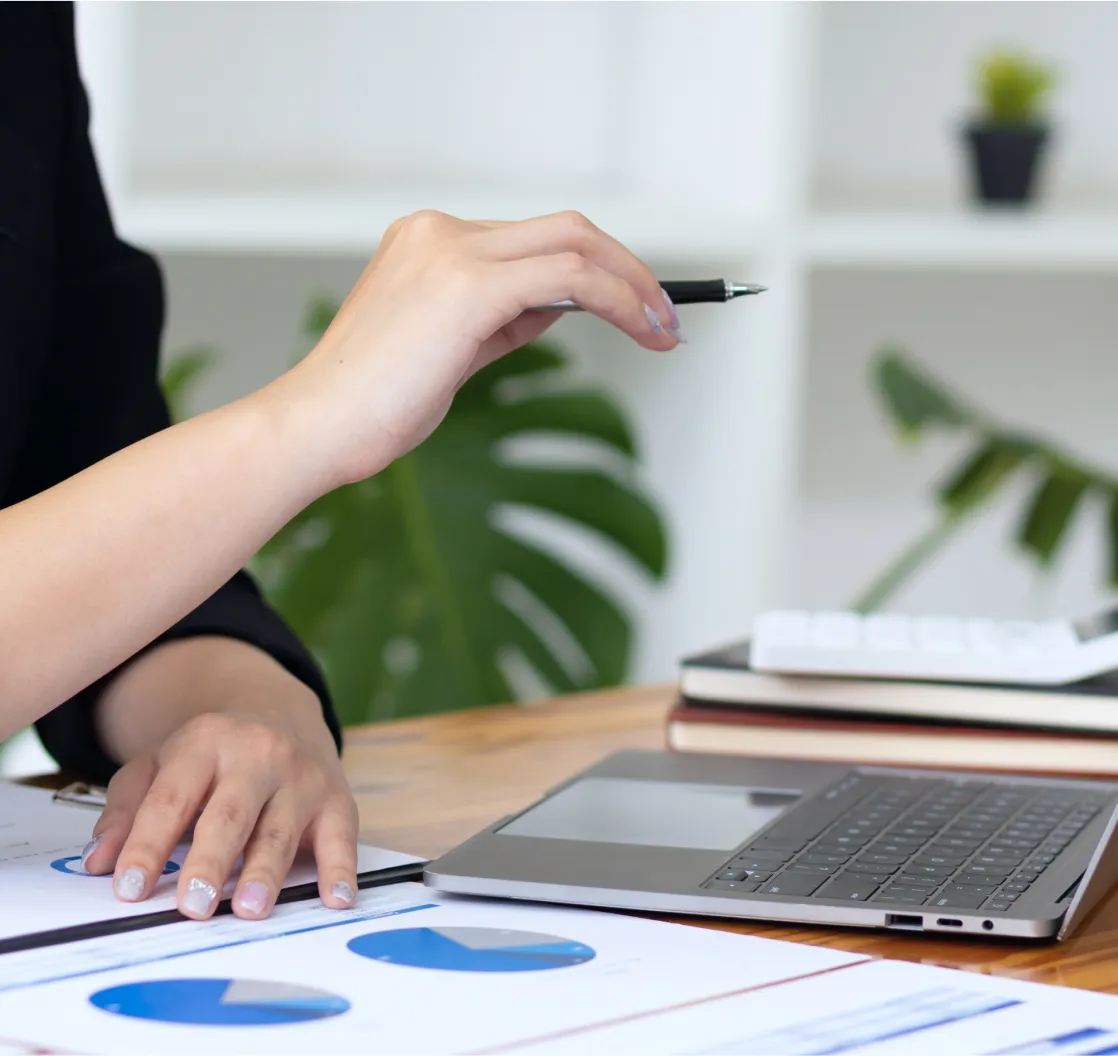 A person holding a pen and pointing at a report with pie charts, with a laptop and a stack of notebooks on the desk.
