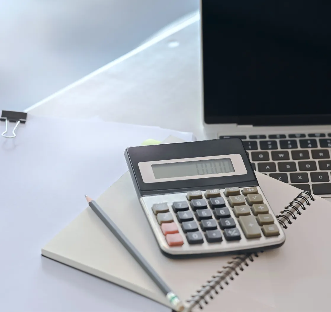 A close-up image of a calculator, pencil, and notebooks placed next to a laptop on a desk.
