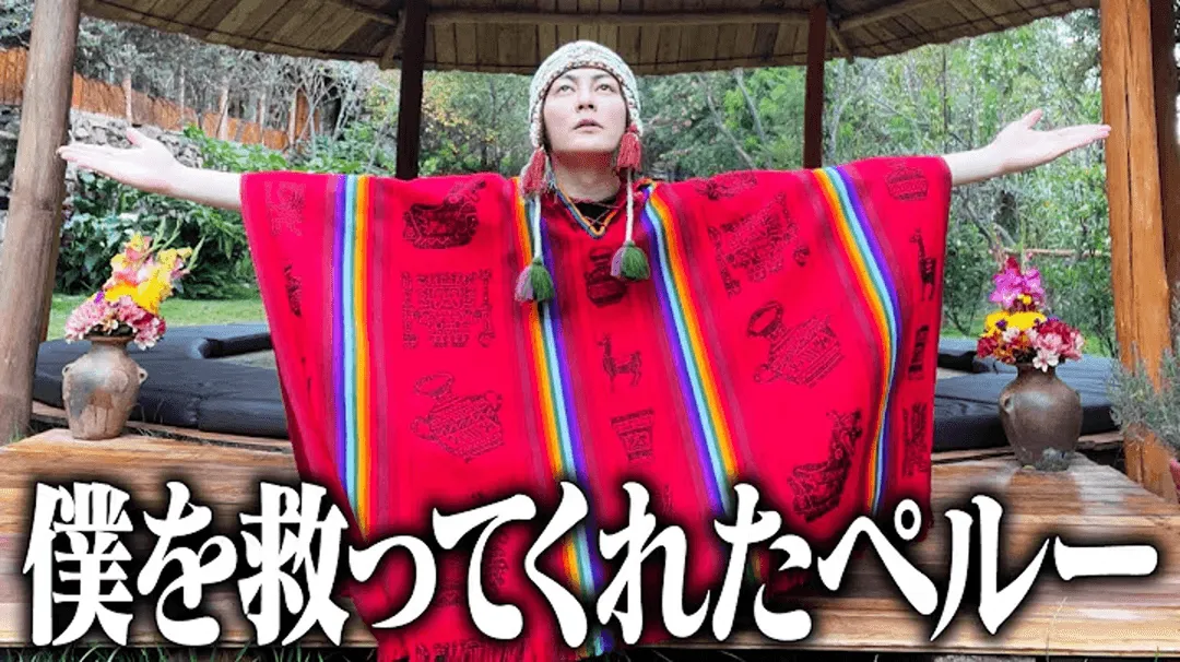 Person wearing a bright red poncho with rainbow stripes and a traditional Peruvian hat, standing with arms wide open under a wooden gazebo surrounded by greenery and flowers.