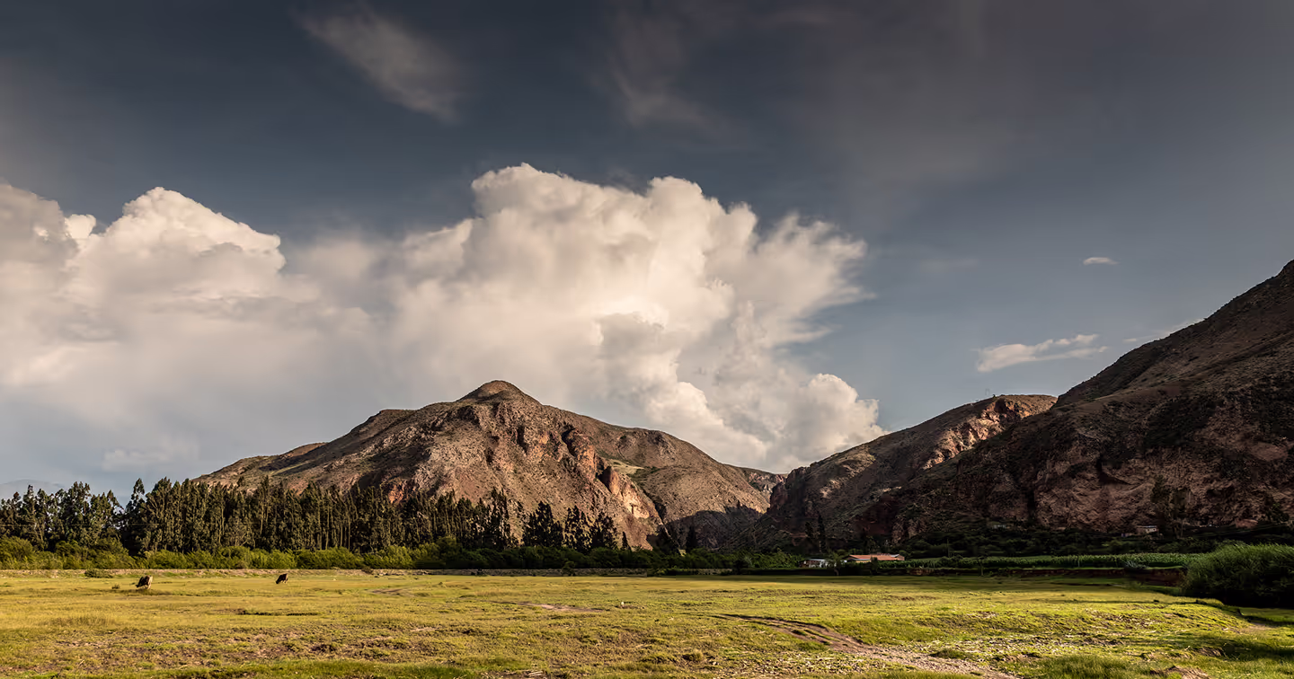 Wide landscape of the Sacred Valley showing grassy fields with grazing animals, surrounded by rugged mountains and a sky filled with dramatic clouds.