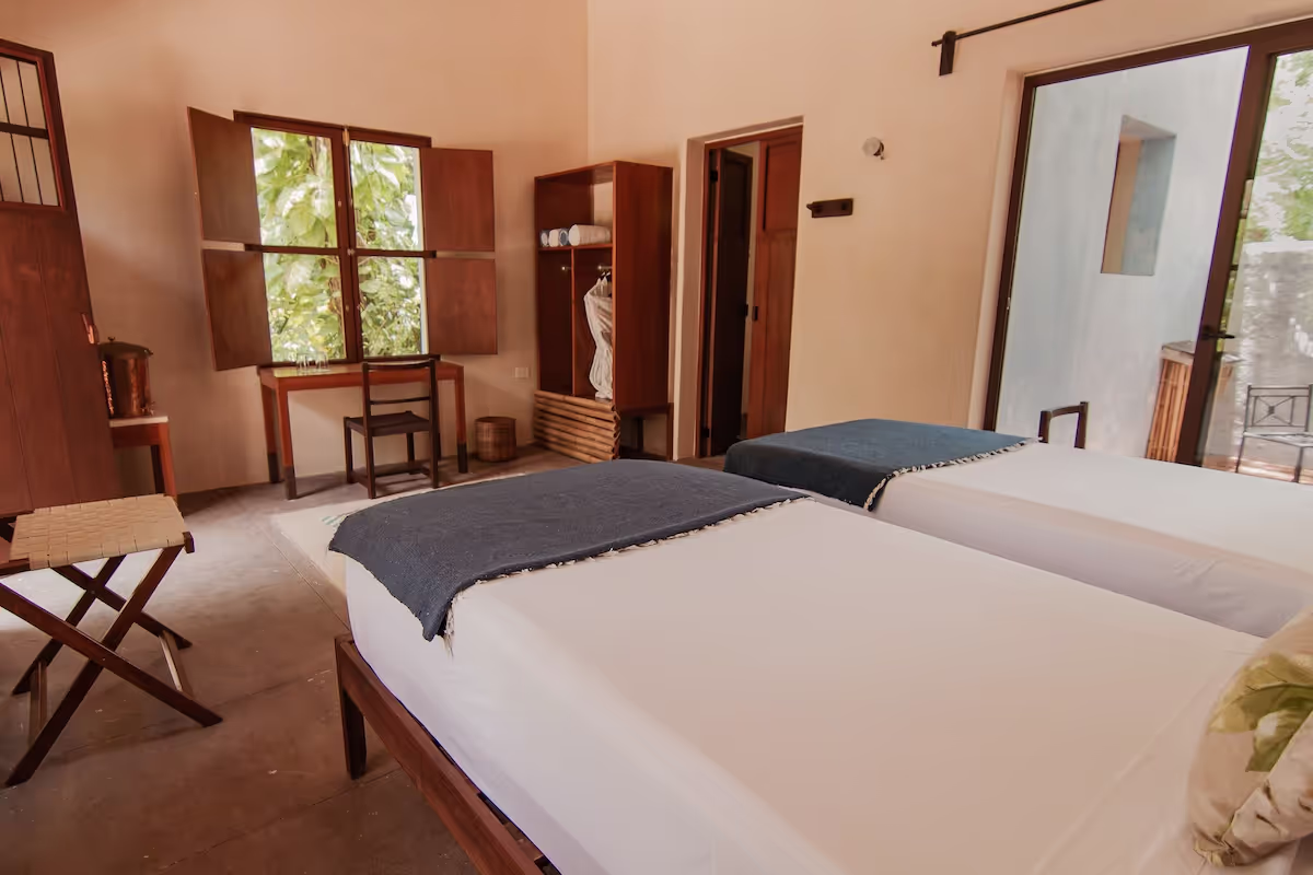 A simple and cozy bedroom featuring two twin beds with white linens and dark blue blankets. The room has wooden furniture including a desk with a chair, an open wardrobe with clothes, and a stool. There is a window with wooden shutters open to reveal greenery outside, and a glass door leading to an outdoor area. The room has a warm, natural ambiance with beige walls and tiled floors.