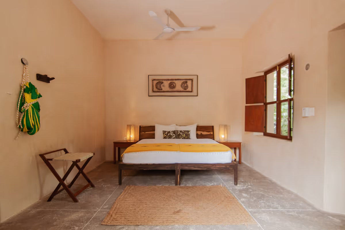 A simple bedroom with beige walls and stone floor. A wooden bed with white bedding and a yellow throw is centered against the back wall, flanked by two wooden nightstands with lit lamps. A small folding stool is to the left, and a window with wooden shutters is open on the right. A woven rug lies on the floor in front of the bed.