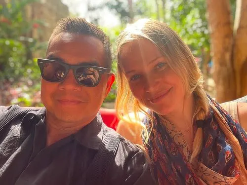 A man wearing black sunglasses and a black shirt sits closely next to a woman with blonde hair styled in a braid. She wears a colorful scarf. Bright sunlight filters through the background, casting a warm glow around them. Both are smiling gently at the camera.