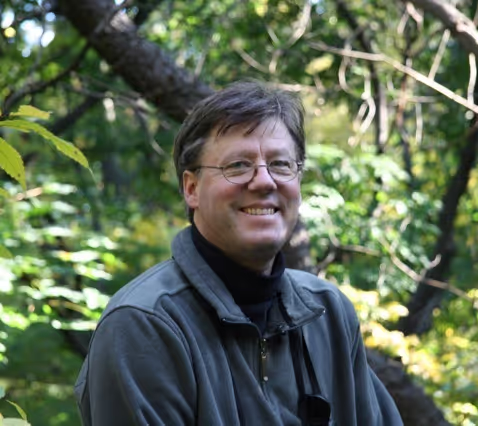 Smiling man wearing a dark blue jacket in the lush green amazon forrest.