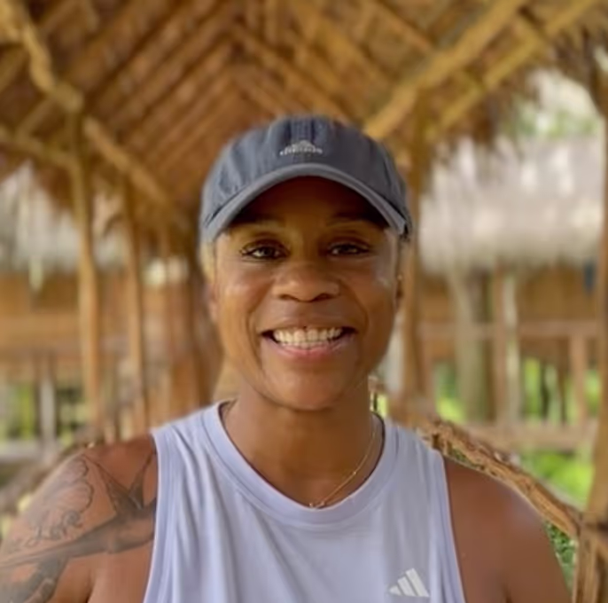 Portrait of a smiling woman with a tattoo on her left shoulder, wearing a black Adidas baseball cap and a white Adidas sleeveless tank top. She stands under a thatched roof structure with wooden supports, with a blurred natural background behind her.