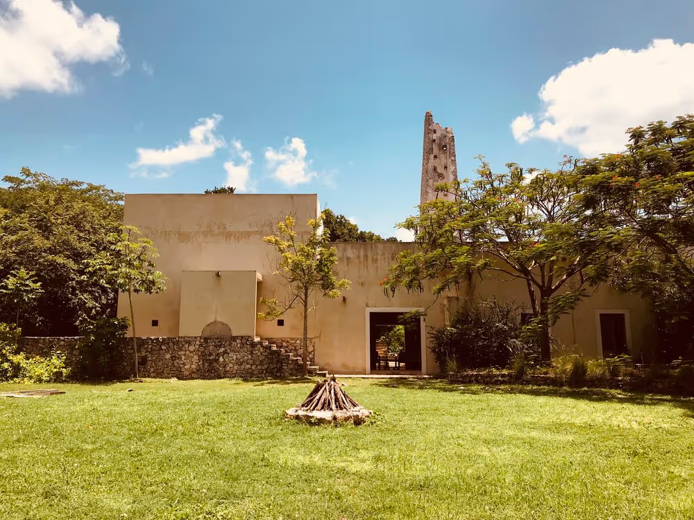 Sunny courtyard Mexican retreat center with green grass, a small campfire setup in the center, surrounded by trees and a beige building with an open doorway. A tall, weathered stone tower rises behind the building under a bright blue sky with scattered clouds.