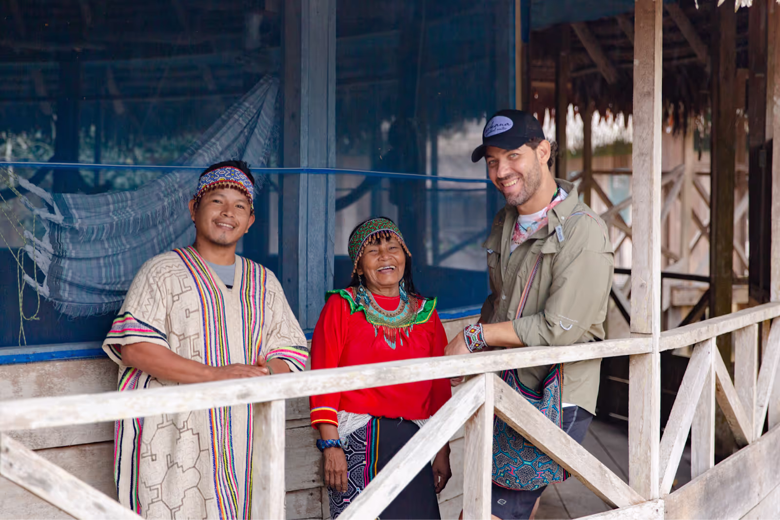 Three smiling people standing on a wooden porch, wearing colorful traditional Amazonian attire with intricate beadwork and patterns, against a backdrop of hammocks and rustic wooden structures.