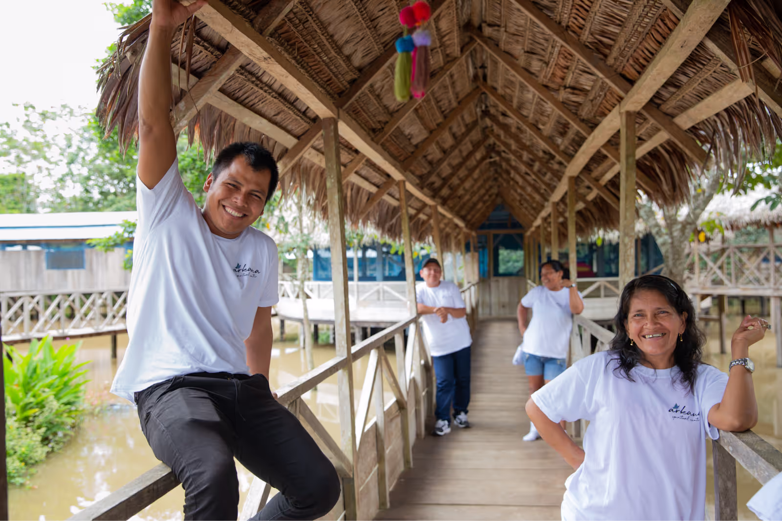 A cheerful group of Arkana Spiritual Center staff wearing matching white t-shirts relax and smile while standing and sitting on a wooden walkway with a thatched roof over water in the Amazon jungle.