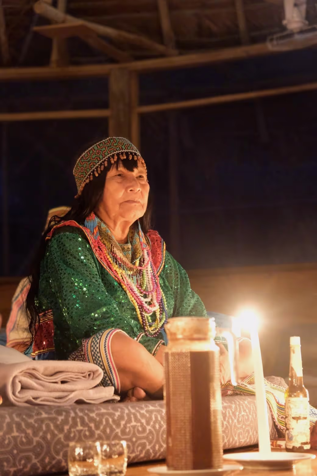 An indigenous shamanic elder wearing a green sequin garment and colorful beaded necklaces and headdress sits solemnly during a candlelit ceremony inside a rustic wooden structure. A jug and candles are on the table before her.