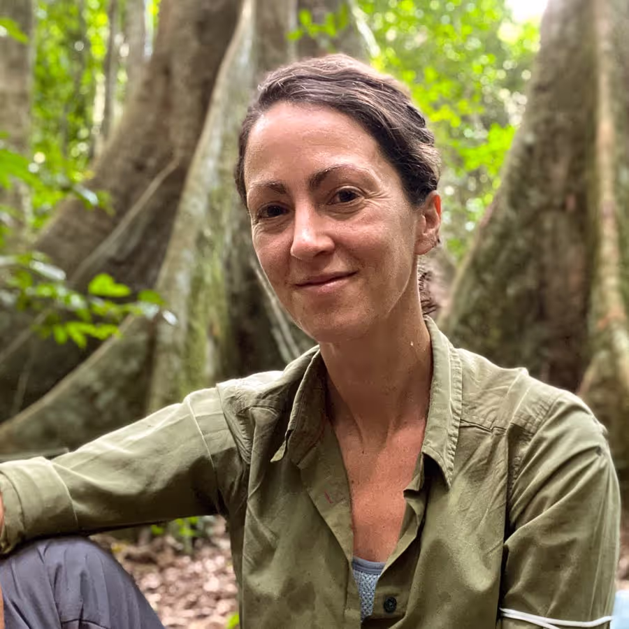 Close-up of a woman with dark hair tied back, wearing a green button-up shirt, sitting outdoors in a dense jungle with large tree roots and green foliage in the background. She smiles gently at the camera.