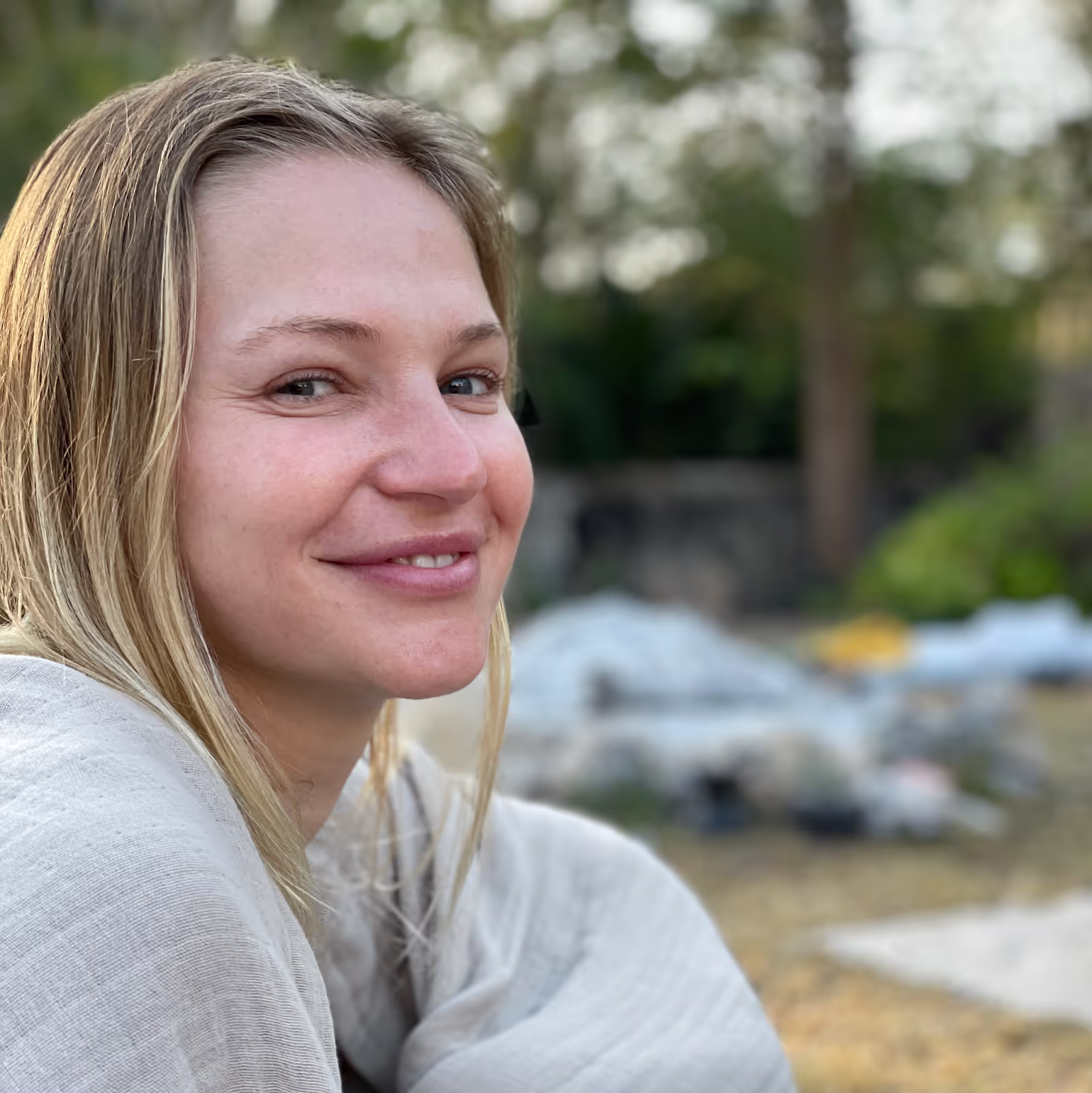 Close-up of a blonde woman with light skin smiling gently, wrapped in a light-colored blanket, sitting outside with blurred greenery in the background.