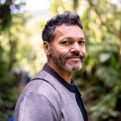 Man with short curly hair and beard looking thoughtfully at camera while wearing a gray blazer over dark shirt, standing in a lush green forest environment with soft natural lighting.