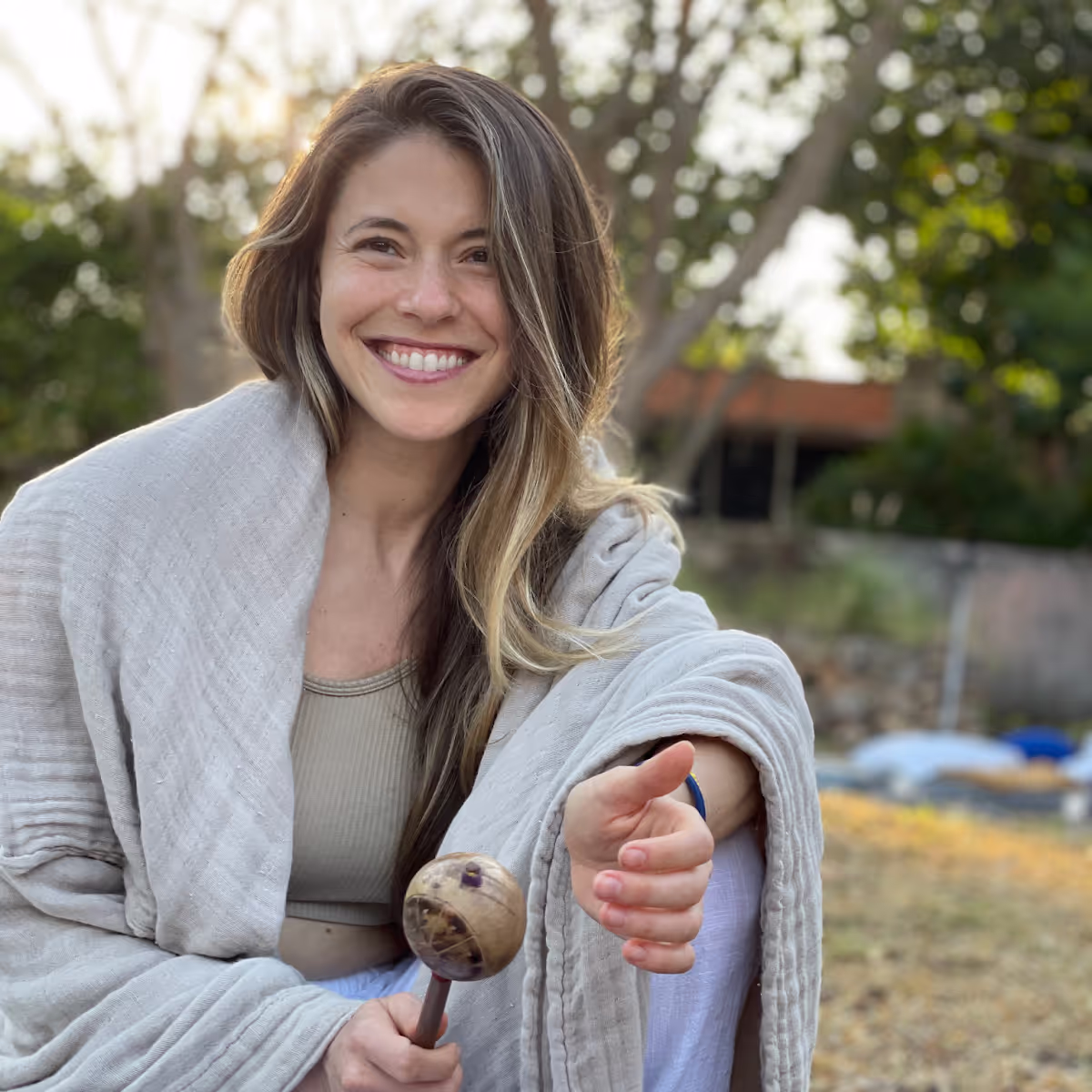 Woman with long hair smiling broadly while wearing light-colored wrap, holding a natural seed rattle or shaker instrument in natural outdoor setting with golden hour lighting.