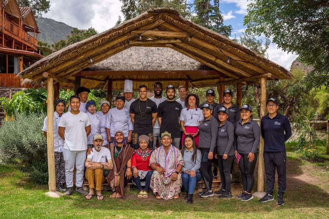 Diverse group of chefs, staff and indigenous community members gathered under a thatched pavilion, with some wearing traditional Andean clothing and others in professional culinary uniforms, surrounded by mountain landscape.