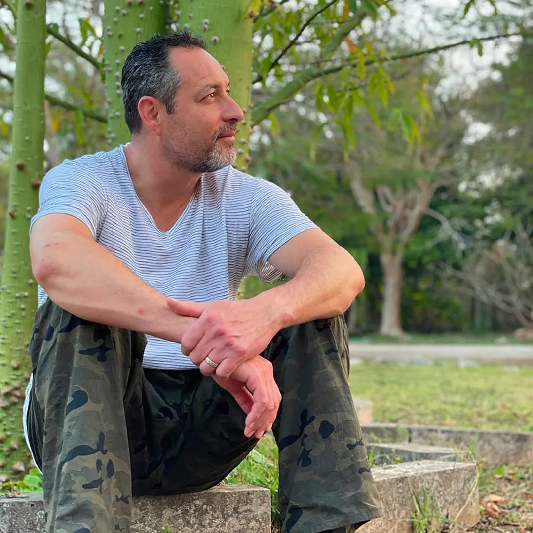 Man in a striped t-shirt and camouflage pants sitting on a low stone ledge next to green tree trunks, with a grassy park and trees in the background on a calm, sunny day.
