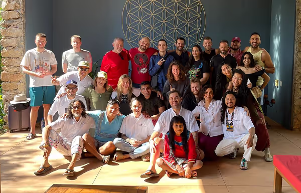 Large group of people posing together in a sunlit lounge area of a retreat center, seated and standing in front of a wall featuring a prominent gold sacred geometry artwork. The space has stone accents, wooden furniture, and a welcoming, communal atmosphere.