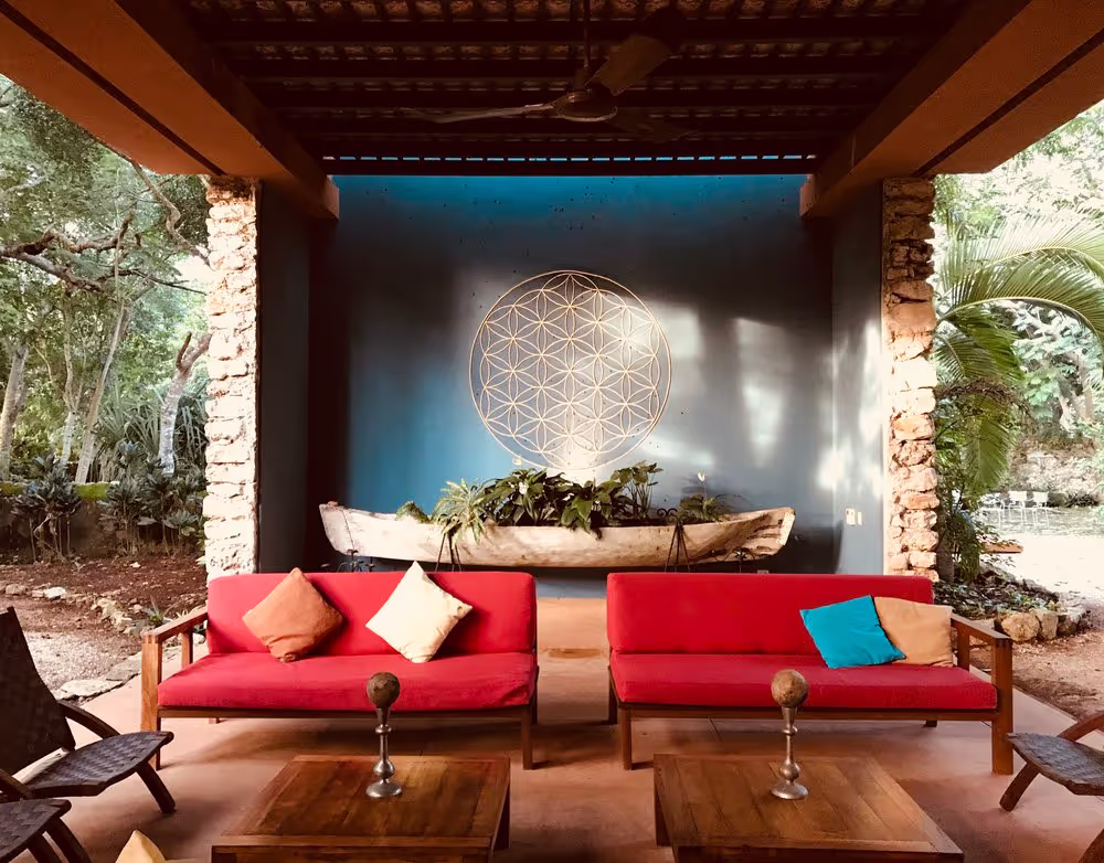 Inviting outdoor lounge area at a Mexican retreat center, featuring two red-cushioned wooden sofas with colorful pillows, a rustic canoe planter filled with greenery, and a large sacred geometry wall art piece on a blue accent wall. The space is framed by stone pillars and surrounded by lush tropical gardens, creating a peaceful and welcoming atmosphere for guests.