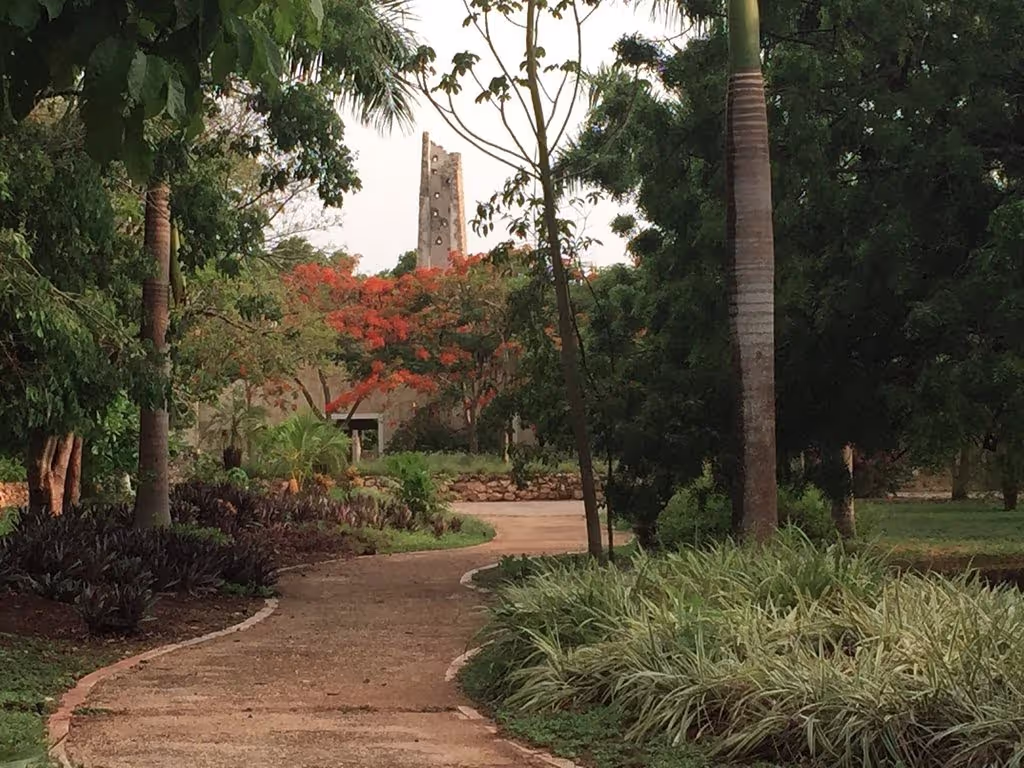 Curved stone pathway leading through vibrant tropical gardens with palm trees and flowering plants, approaching a retreat center building with a unique tower structure in the background. The scene is tranquil, inviting visitors to explore the peaceful, green retreat grounds.