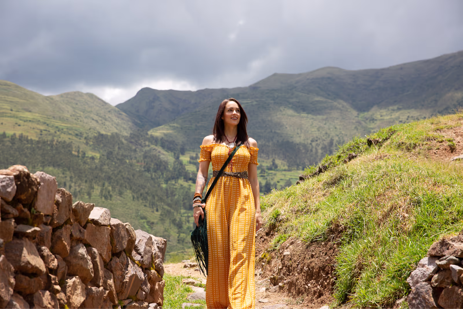 Woman wearing a yellow off-shoulder dress walking along a rustic stone path in a lush green mountainous landscape under a cloudy sky.