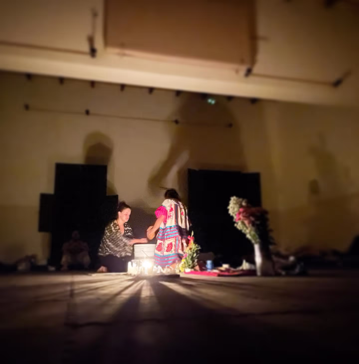 A dimly lit ceremonial space where a facilitator in traditional Shipibo clothing conducts a personal healing session with a participant, surrounded by candlelight that creates atmospheric shadows on the walls.