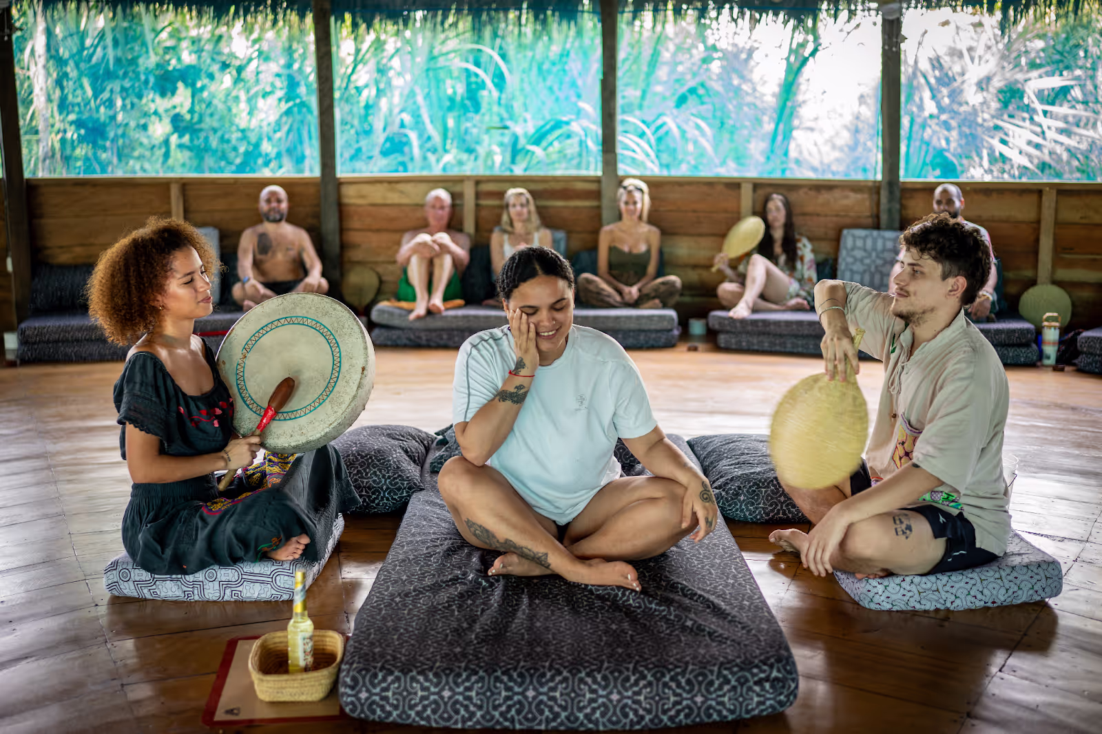 Three facilitators with traditional drums lead a healing ceremony in a wooden maloca with jungle views, while retreat participants sit on meditation cushions in a sacred circle formation.