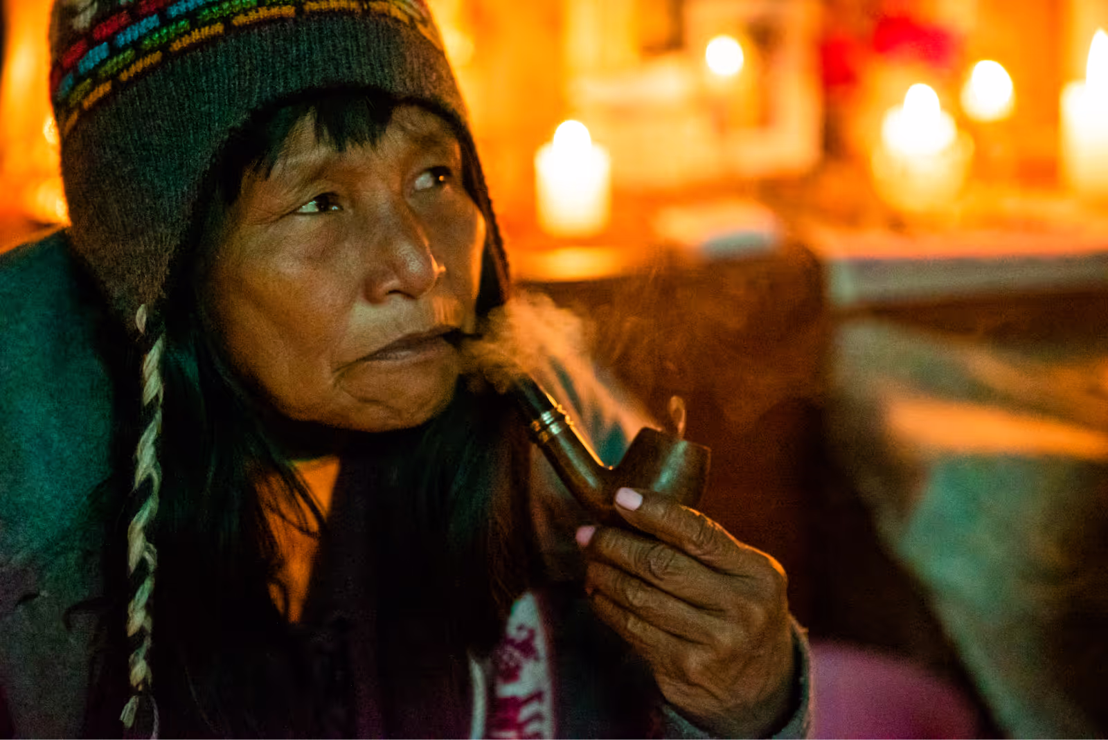 Close-up of a shaman wearing a colorful knit hat and traditional clothing, smoking a wooden pipe inside a dimly lit room illuminated by candles, with smoke visible in the air.