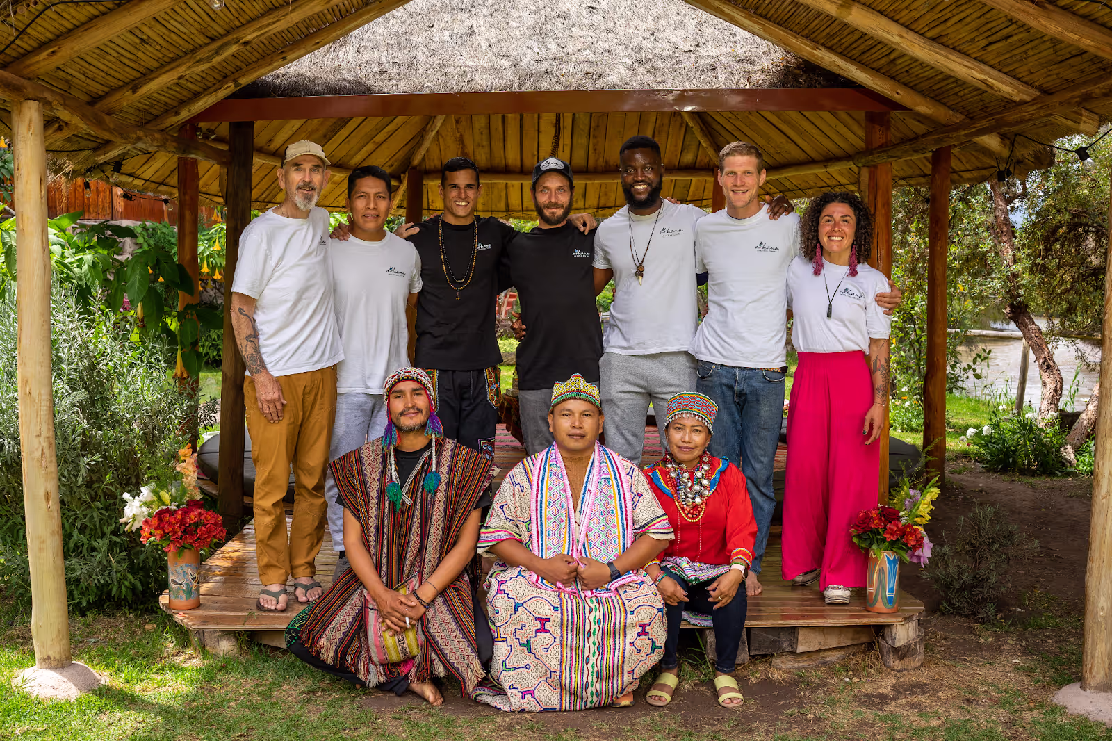 A group photo of Arkana Ayahuasca Retreat staff and shamans. Seven casually dressed staff members stand in the back row wearing Arkana t-shirts. Three shamans in colorful traditional clothing and hats sit cross-legged in front on a wooden platform surrounded by greenery.